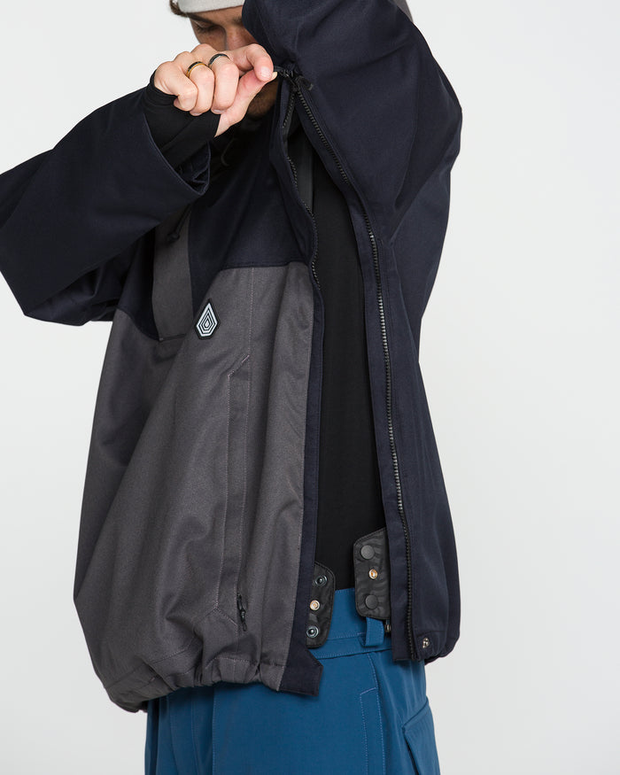 A person wears the Volcom Men’s Hossegor 20K Pullover—a dark recycled V-Science shell jacket—paired with blue pants, raising their right arm to zip or adjust the jacket and partially covering their face, against a plain white background.