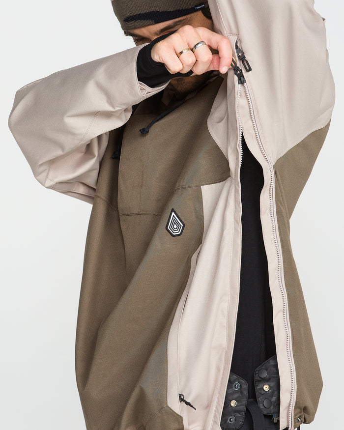 A person in a brown and beige Men’s Hossegor 20K Pullover by Volcom is zipping it up, their face partially obscured by the arm and hood. This pullover features a geometric chest patch and is crafted from recycled V-Science shell material for enhanced sustainability.