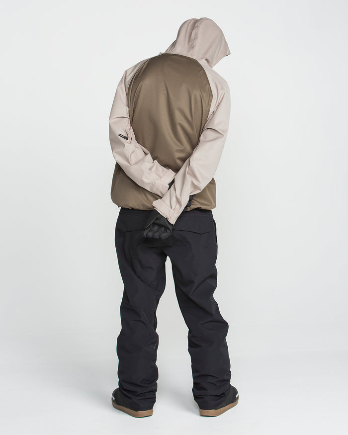 A person wearing the Volcom Men’s Hossegor 20K Pullover—a tan and brown hooded jacket crafted from recycled V-Science shell with a PFAS-free DWR finish for added protection—paired with black pants stands with their back to the camera, head tilted down, and hands clasped behind their back against a plain white background.
