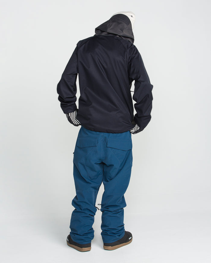 A person stands facing away against a plain white background, wearing the Volcom Men’s Hossegor 20K Pullover—a black jacket crafted from recycled V-Science shell—paired with blue snow pants, striped gloves, and a white beanie.