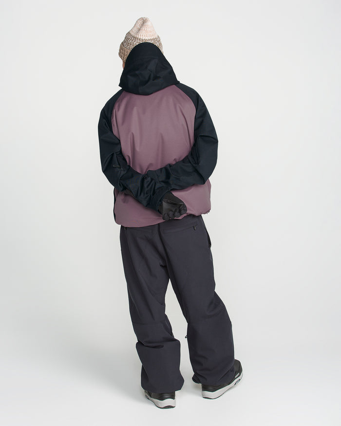 A person stands with their back to the camera, arms crossed behind them, wearing the Volcom Men’s Hossegor 20K Pullover in pink and black. The jacket is crafted from a recycled V-Science shell and features PFAS-free DWR for eco-friendly water resistance. They are also wearing black pants, black gloves, gray shoes, and a beige knit hat, all set against a plain white background.
