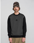 A man wearing the Volcom Men’s Hydro Crew, a dark gray and black sweatshirt made from hydrophobic fleece for moisture-wicking comfort and dryness all day, pairs it with beige pants and a brown patterned beanie while standing against a plain light gray background and looking at the camera with a neutral expression.