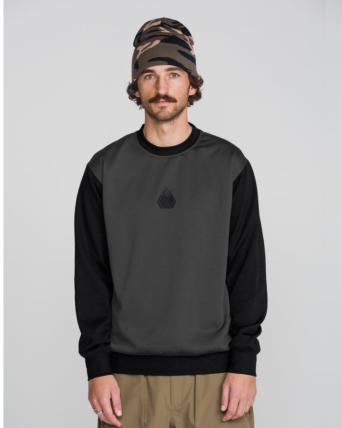 A man wearing the Volcom Men’s Hydro Crew, a dark gray and black sweatshirt made from hydrophobic fleece for moisture-wicking comfort and dryness all day, pairs it with beige pants and a brown patterned beanie while standing against a plain light gray background and looking at the camera with a neutral expression.