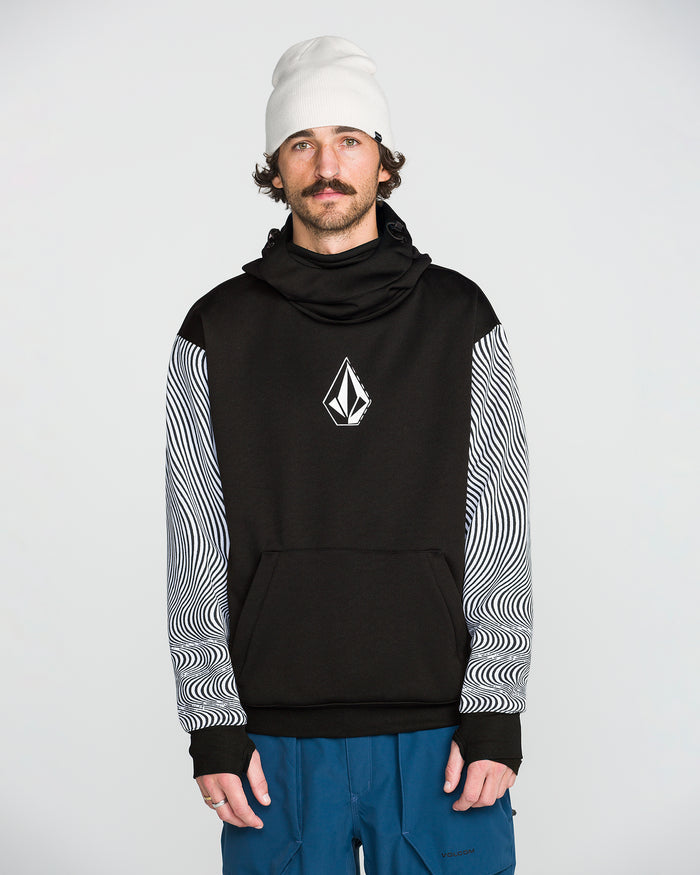 A man wearing the Volcom Men’s Hydro Riding Hoodie featuring a geometric Volcom Stone logo and patterned sleeves, paired with a white beanie and blue pants, stands against a plain light background.