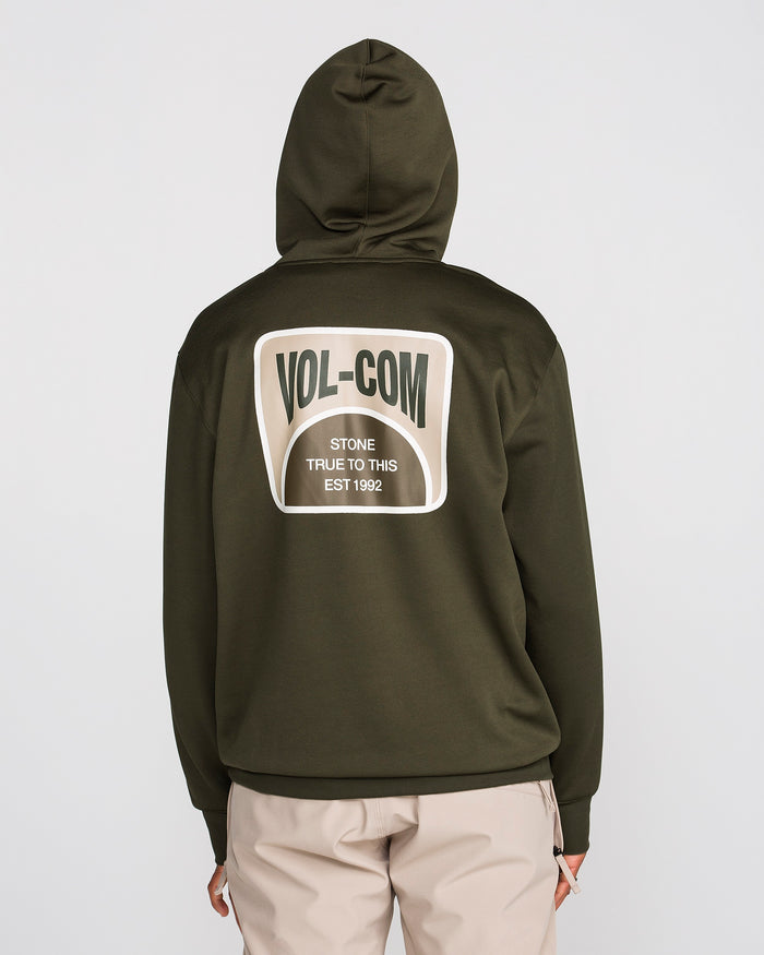A person is wearing a Volcom Men’s Hydro Riding Hoodie in olive green with the hood up. The back of the hoodie features a large patch that reads “VOL-COM Stone True To This EST 1992.” They are also wearing beige pants.