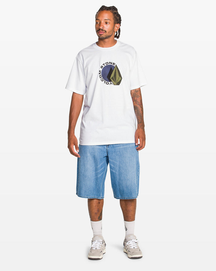 A man in a white Volcom Men's Hypnotizer Short Sleeve Tee, light blue knee-length denim shorts, white socks, and beige sneakers stands against a plain white background.