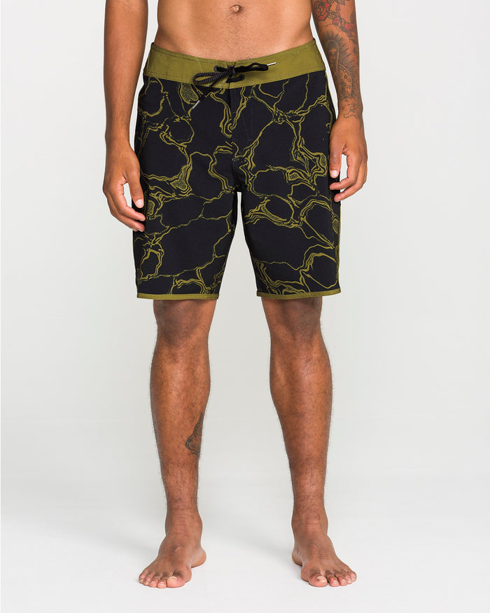 A person stands barefoot against a plain white background, hands relaxed at their sides, wearing the Volcom Men's Infuse Scallop Mod boardshorts in black and olive-green with a yellow marble pattern and 4-way stretch for enhanced comfort.