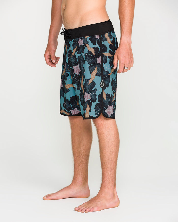 A person stands barefoot on a white background, wearing Volcom Men's Infuse Scallop Mod board shorts featuring a teal and black pattern, drawstring waist, and 4-way stretch for maximum mobility. Only the lower half of the body is visible.