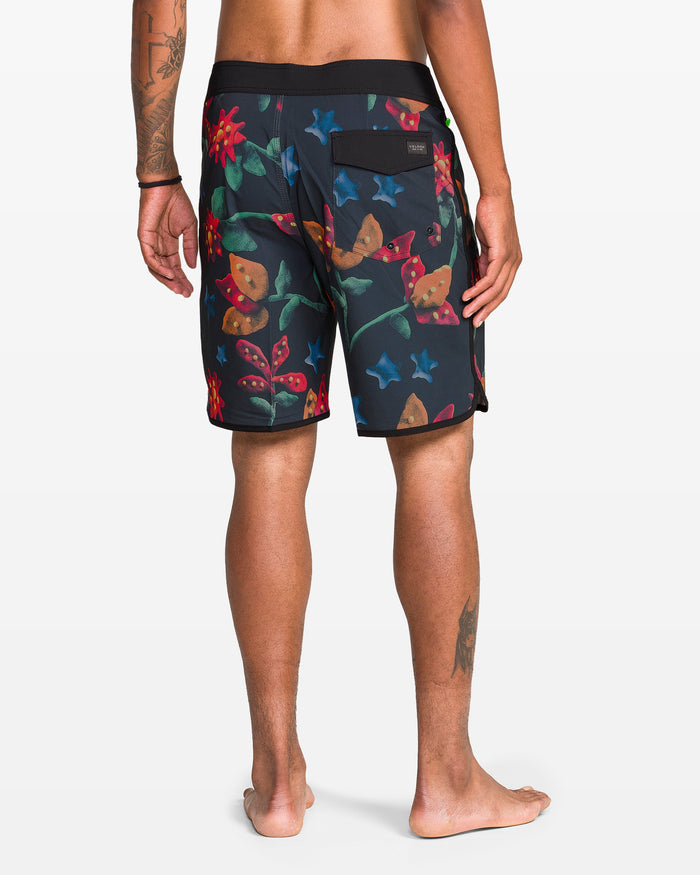A person stands barefoot, facing away, wearing Volcom Men's Infuse Scallop Mod Boardshorts with a dark floral pattern of butterflies, red flowers, green leaves, and a back pocket. Their left arm and right calf tattoos are visible.