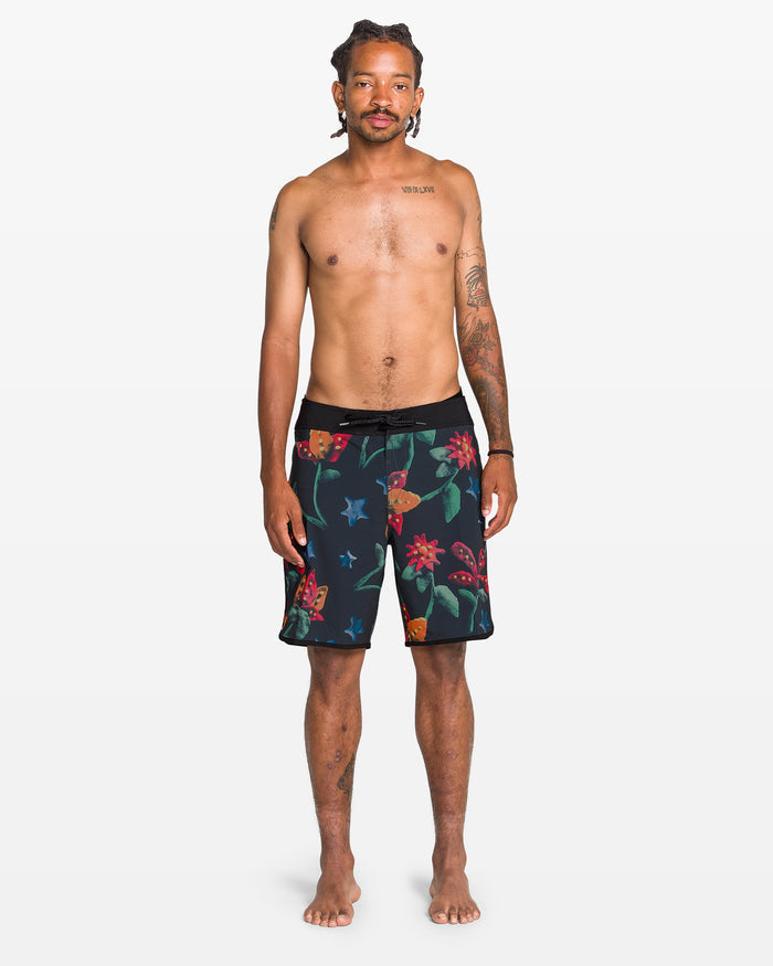 A tattooed man stands barefoot against a white background, facing forward in Volcom Men's Infuse Scallop Mod Boardshorts, which feature a colorful floral pattern and Cinch Fly Technology. He is shirtless with a neutral expression.