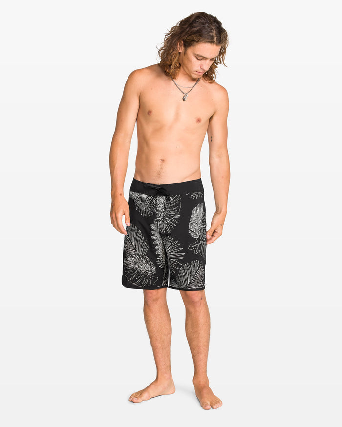 A barefoot man with long hair, a necklace, and black Volcom Men's Infuse Scallop Mod boardshorts featuring a white tropical leaf pattern and Cinch Fly Technology stands against a plain white background, looking down.