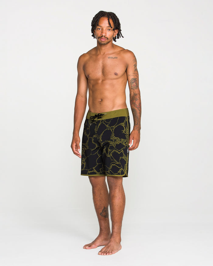 A man with short dreadlocks and tattoos stands barefoot, wearing Volcom Men's Infuse Scallop Mod boardshorts in black and olive green floral 4-way stretch fabric with a Cinch Fly, against a plain white background.
