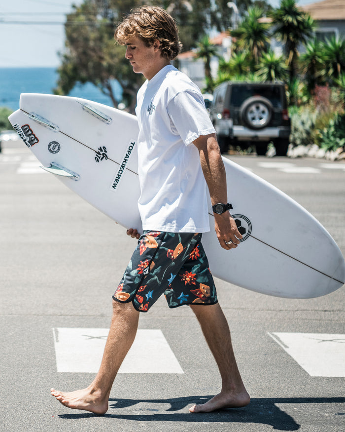 A young man wearing Volcom's Men's Infuse Scallop Mod boardshorts and a white t-shirt walks barefoot across a street with a surfboard, while palm trees, parked cars, and the ocean appear in the background.