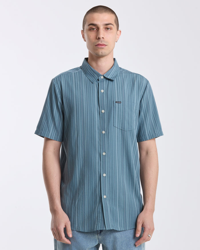 A person with short hair wears the Volcom Men's Jahstripe Short Sleeve Shirt in blue, paired with light blue jeans, standing against a plain white background.