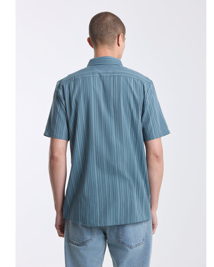 A person with short hair is seen from behind, wearing a blue Volcom Men's Jahstripe Short Sleeve Shirt and light blue jeans against a plain white background.