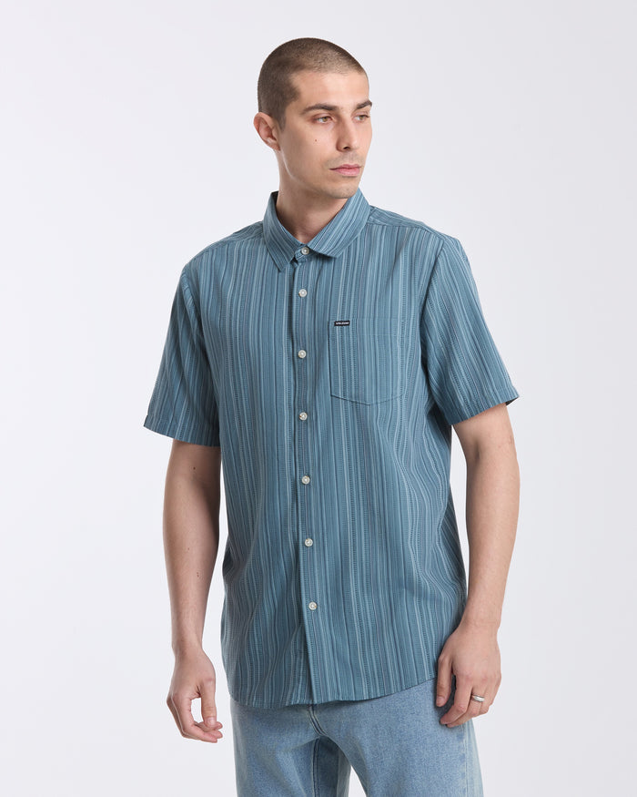 A man with short brown hair wears the Volcom Men's Jahstripe Short Sleeve Shirt—a blue, classic fit shirt with vertical stripes—and light blue jeans, standing against a plain light gray background and looking slightly to his left.