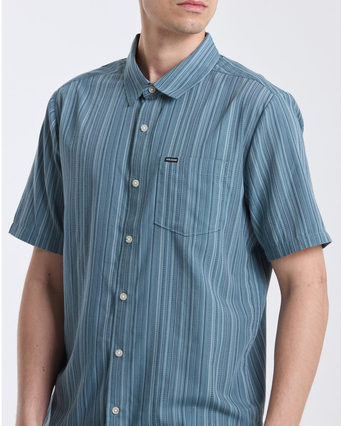 A person wearing the Volcom Men's Jahstripe Short Sleeve Shirt, featuring a blue pattern and front pocket, stands against a plain light background.