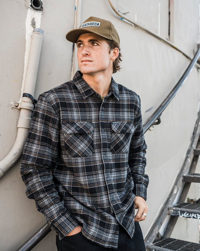 Wearing the Volcom Men’s Johnstone Long Sleeve Shirt, a man with a relaxed, boxy fit and tan cap leans against a wall lined with pipes, gazing to the side with his hands in his pockets and stairs behind him.