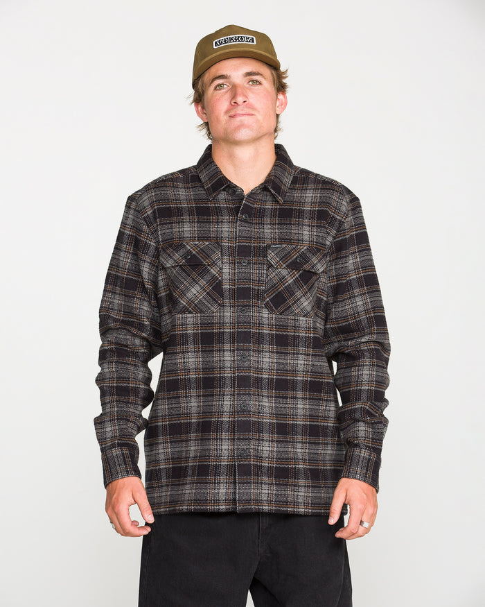 A person wearing a beige cap, black pants, and the Volcom Men's Johnstone Long Sleeve Shirt — a black-and-brown plaid heavyweight flannel with chest pockets — stands facing forward against a plain white background.