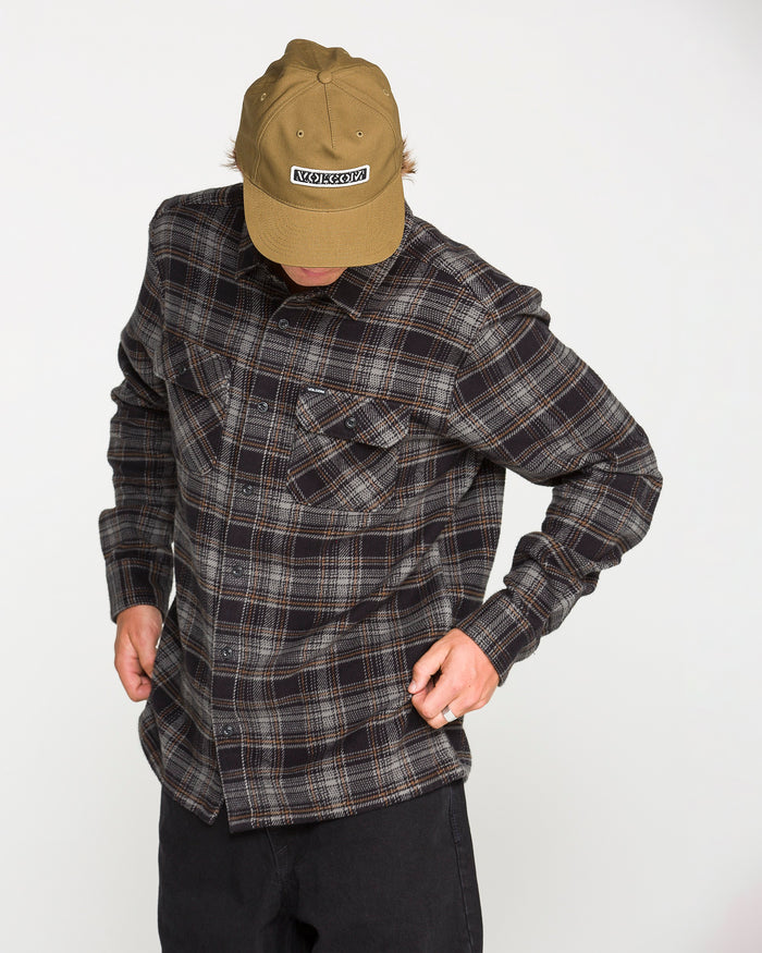 A person wearing a tan cap, the Volcom Men's Johnstone Long Sleeve Shirt in a dark color, and black pants looks down while adjusting their shirt against a plain white background.