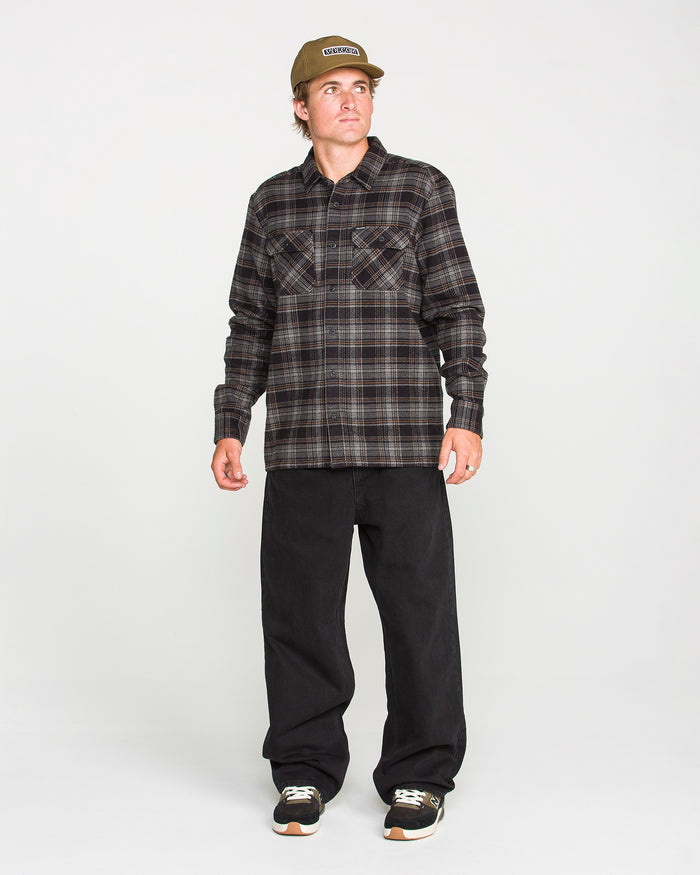 A person wearing the Volcom Men's Johnstone Long Sleeve Shirt in brown plaid, paired with black pants, black sneakers, and a tan cap stands against a plain white background, looking slightly to their left. The shirt's brushed flannel fabric adds texture and warmth to their casual look.