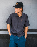A young man, wearing the Volcom Men’s Kap Out Workwear Short Sleeve Shirt—featuring a dark, durable design with chest pockets—and blue jeans, leans against a gray wall. He completes the look with a black cap, gazing to his left with his hands in his pockets.