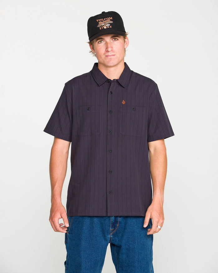 A person wearing the Volcom Men's Kap Out Workwear Short Sleeve Shirt in black, paired with blue jeans and a black cap featuring orange text, stands against a plain white background facing the camera with a neutral expression.