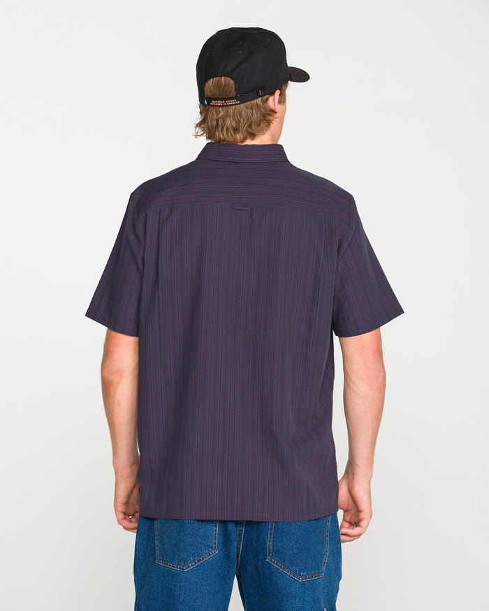 A person is shown from the back, standing against a plain light background, wearing a black cap, blue jeans, and the Volcom Men's Kap Out Workwear Short Sleeve Shirt—a dark button-up with thin stripes.