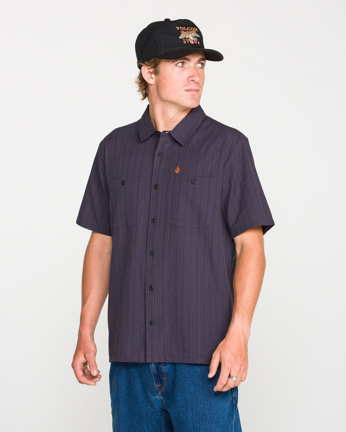 A person wearing a black cap and the Volcom Men's Kap Out Workwear Short Sleeve Shirt—a dark button-up with thin stripes—paired with blue jeans, stands facing left against a plain background.