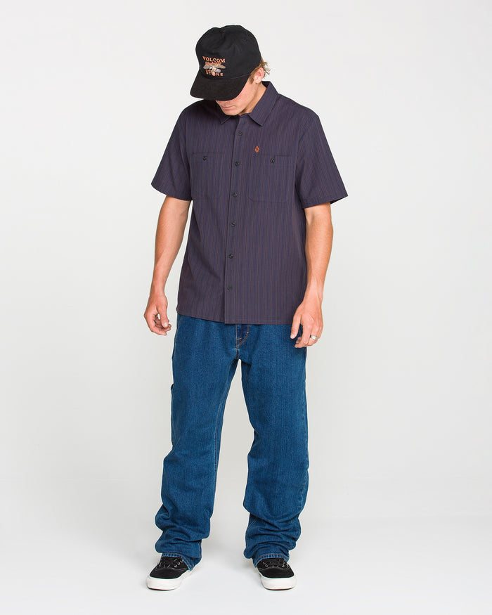 A person wearing a black cap, the Volcom Men's Kap Out Workwear Short Sleeve Shirt, blue jeans, and black sneakers stands against a plain white background, looking down with hands relaxed at their sides.