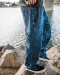 A person wearing Volcom Men’s Kinkade Utility Denim Pants and black sneakers stands on rocks by a body of water, with part of a bridge and trees visible in the background.