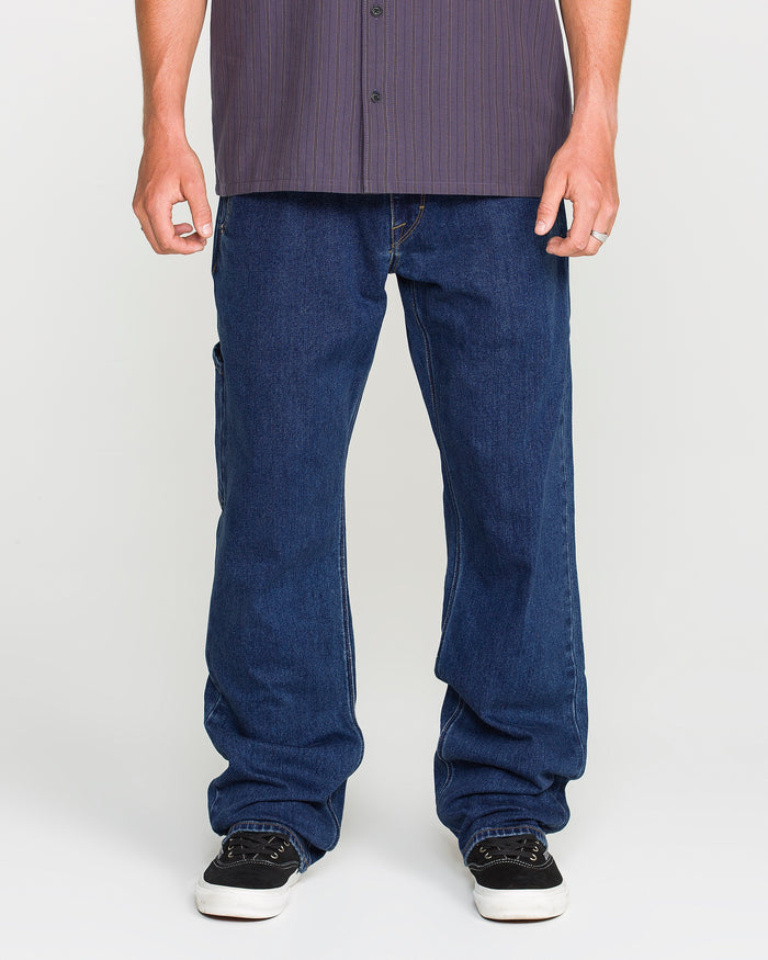A person wearing a dark striped shirt, Volcom Men's Kinkade Utility Denim Pants in indigo, and black shoes stands against a plain white background. Their face is not visible in the image.