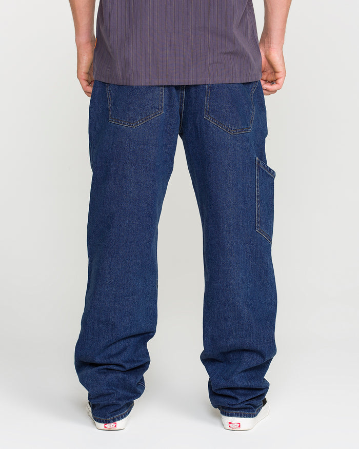 A person wearing Volcom Men's Kinkade Utility Denim Pants in indigo and white sneakers is standing facing away from the camera, with only their lower back, legs, and feet visible.