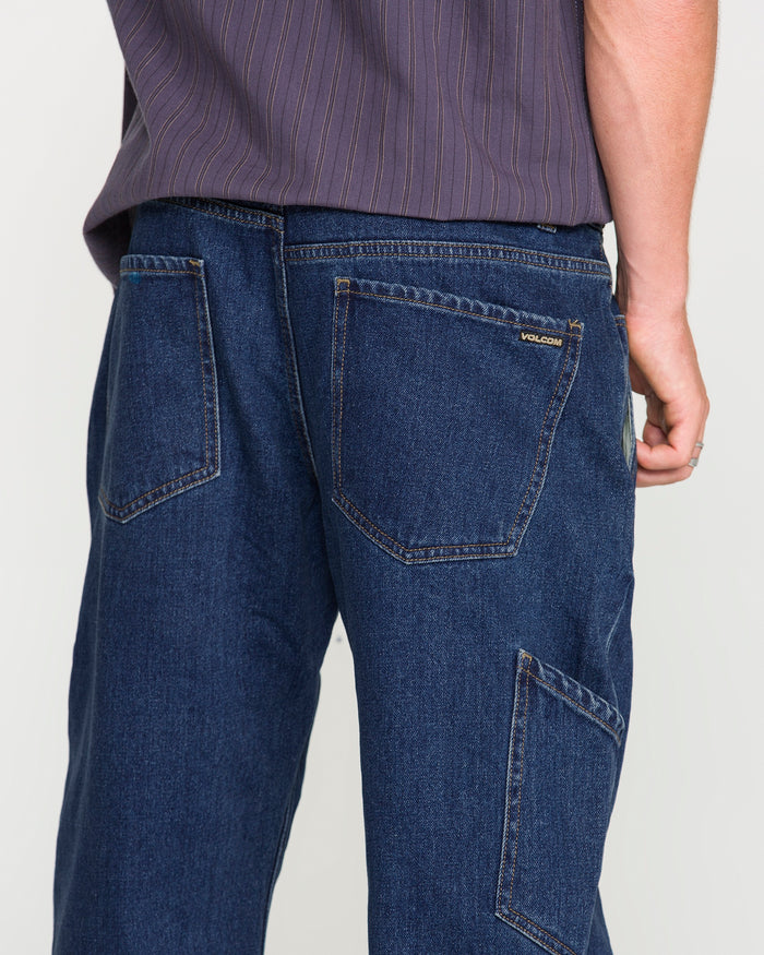 A person wearing Volcom Men's Kinkade Utility Denim Pants in dark blue indigo, featuring visible back and side pockets, paired with a purple striped shirt. The photo is taken from behind, highlighting the straight-leg utility denim pants and part of the shirt.
