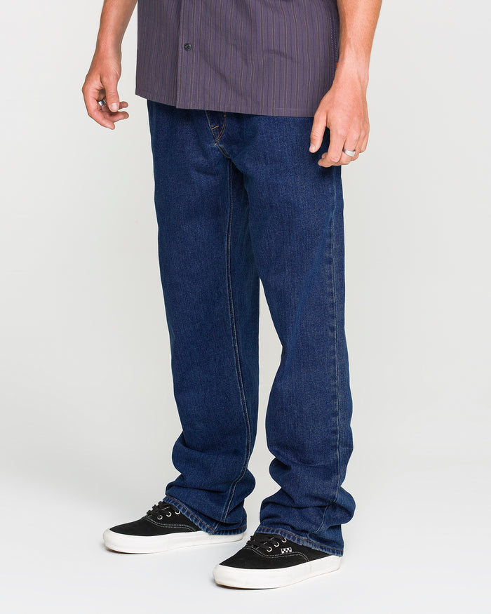 A person wearing a short-sleeved, dark striped shirt, Volcom Men's Kinkade Utility Denim Pants in an indigo wash, and black sneakers with white soles stands against a plain white background.