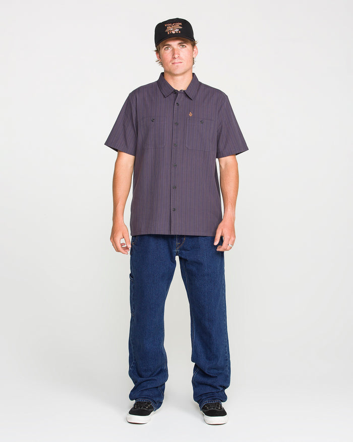 A person in a black cap, short-sleeve button-up shirt, and black shoes stands facing forward against a plain white background, wearing Volcom Men's Kinkade Utility Denim Pants for a timeless look.