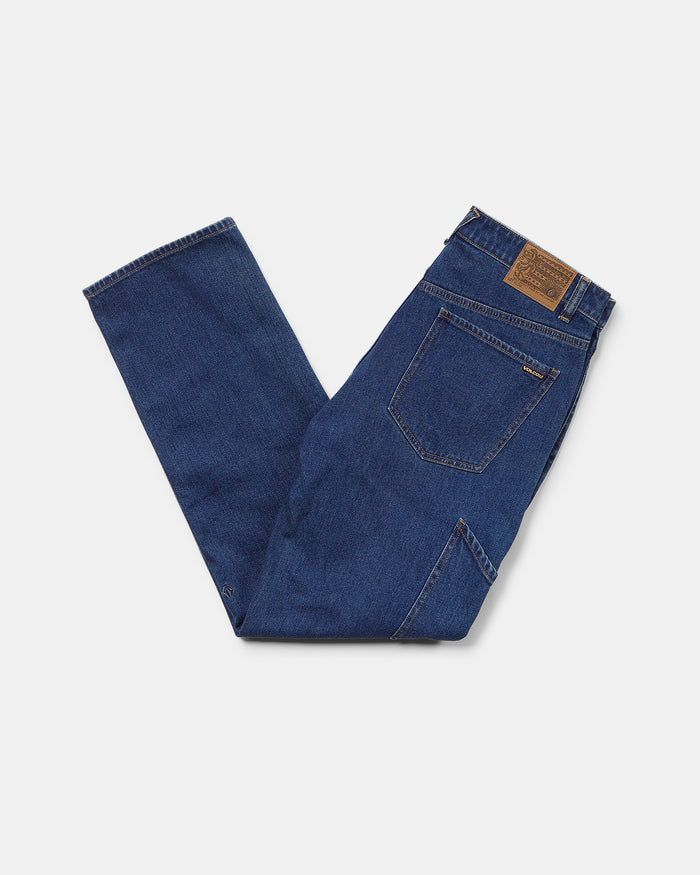 A pair of folded Volcom Men's Kinkade Utility Denim Pants in indigo, featuring visible back pockets and a brown leather patch on the waistband, displayed on a white background.