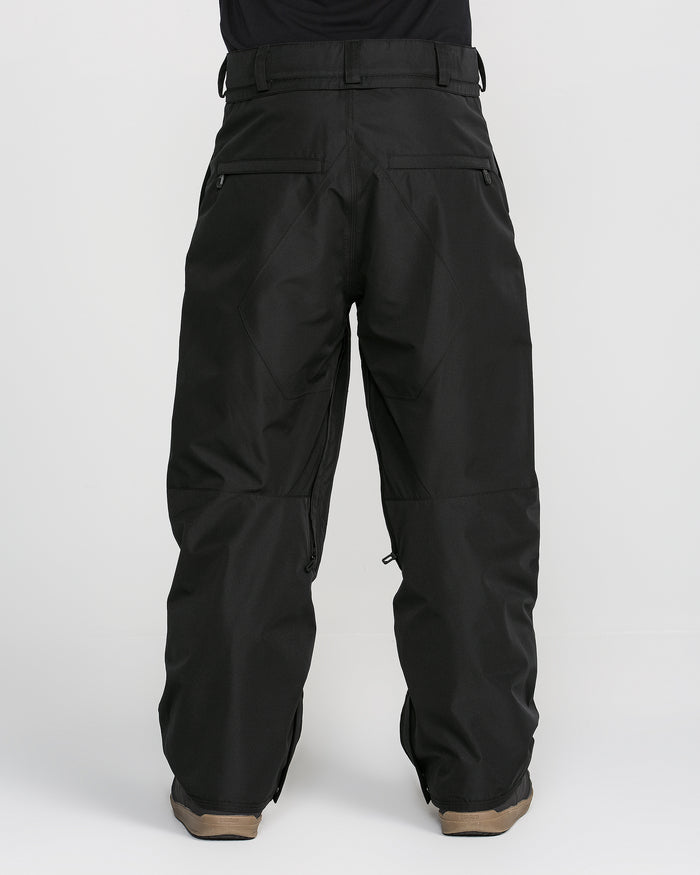 A person is shown from the back wearing loose-fitting black Volcom Men’s Kleveland Pants, with visible pockets and belt loops; these snow pants, featuring V-Science 2-Layer construction, cover brown shoes against a plain white background.