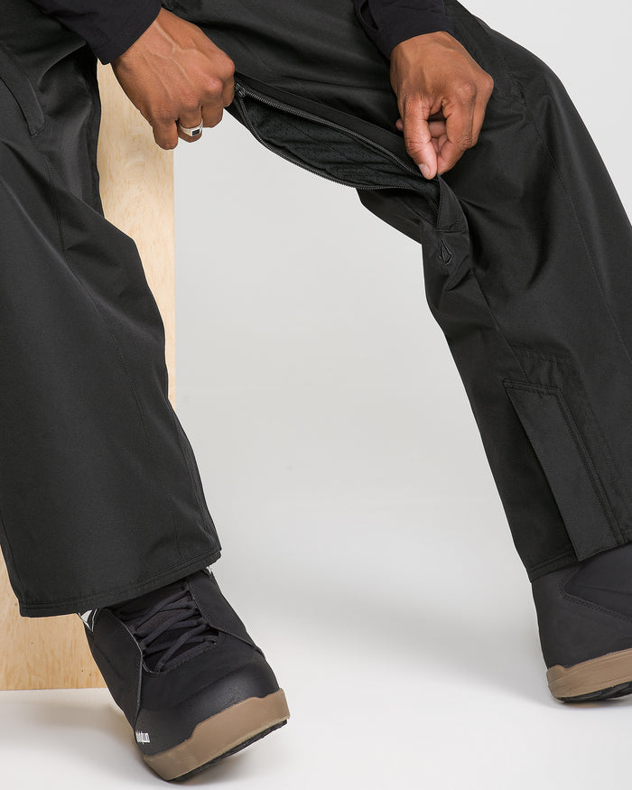 A person is sitting and unzipping a side vent on black Volcom Men’s Kleveland Pants, wearing black shoes with brown soles, against a plain background.