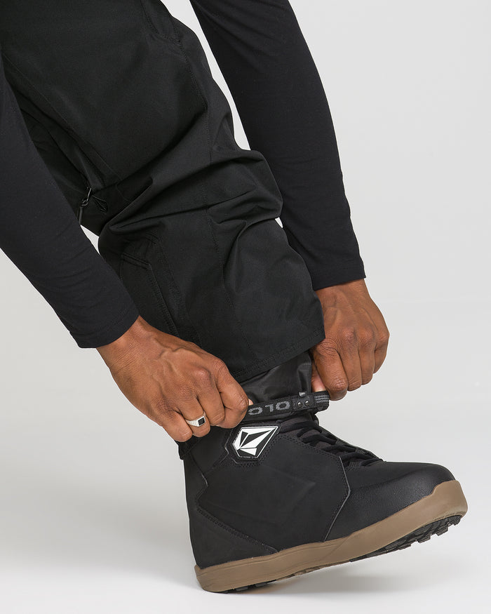 A person wearing black Volcom Men’s Kleveland Pants secures the strap on a black snowboarding boot with a brown sole.
