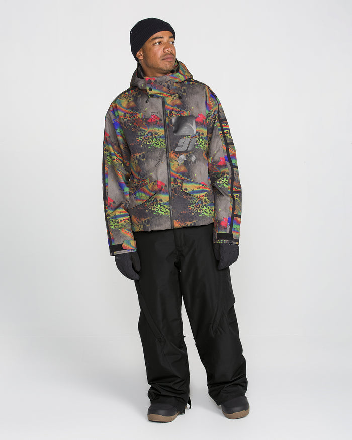 A man wearing a colorful, patterned hooded jacket, black gloves, a black beanie, and Volcom Men’s Kleveland Pants stands against a plain white background, looking slightly to his left.