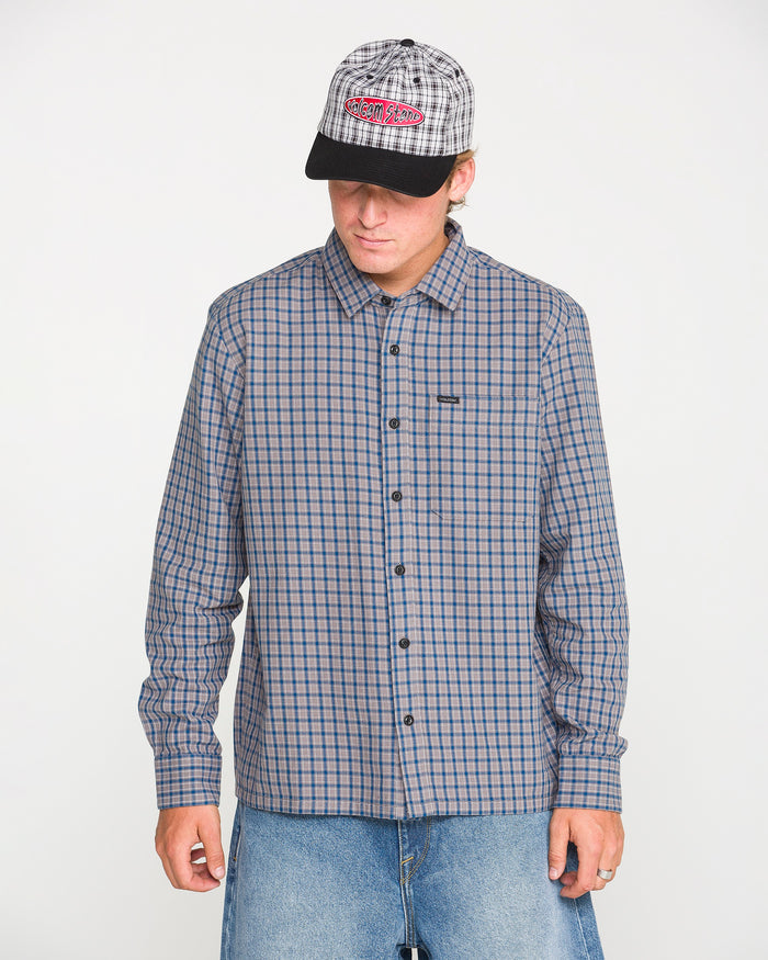 A person wearing the Volcom Men's Landos Long Sleeve Shirt, featuring a checked pattern and an oversized chest pocket, stands against a plain white background looking down. They pair the shirt with light-colored jeans, a plaid baseball cap, and have their hands at their sides.