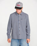 A person wearing the Volcom Men's Landos Long Sleeve Shirt, featuring a checked pattern and an oversized chest pocket, stands against a plain white background looking down. They pair the shirt with light-colored jeans, a plaid baseball cap, and have their hands at their sides.