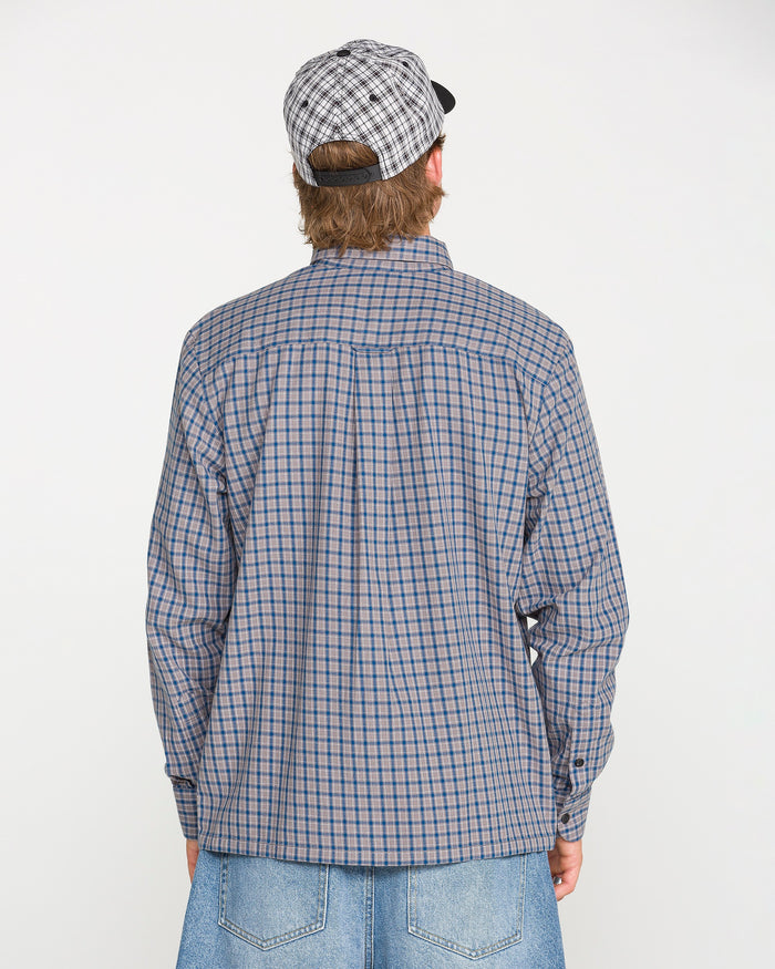 A person is shown from the back against a plain white background, wearing the Volcom Men's Landos Long Sleeve Shirt with a yarn-dyed plaid pattern, a plaid cap, and blue jeans.