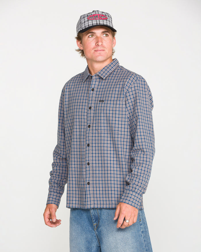 A man wearing the Volcom Men's Landos Long Sleeve Shirt—a gray yarn-dyed plaid button-up with a relaxed, boxy silhouette—paired with blue jeans and a gray baseball cap, stands against a plain white background, looking slightly to the side.