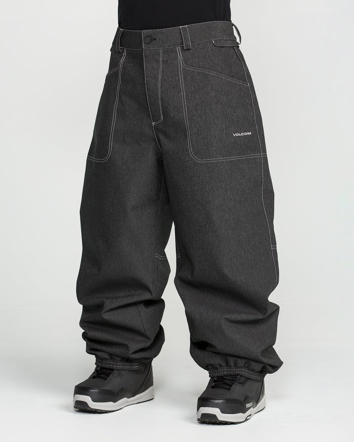 A person is wearing Volcom Men’s Lapper Pants in a loose-fitting, dark gray style with white stitching and gathered hems at the ankles. The pants feature the Volcom logo on the pocket and are paired with a black shirt and black and gray sneakers. The background is plain white.