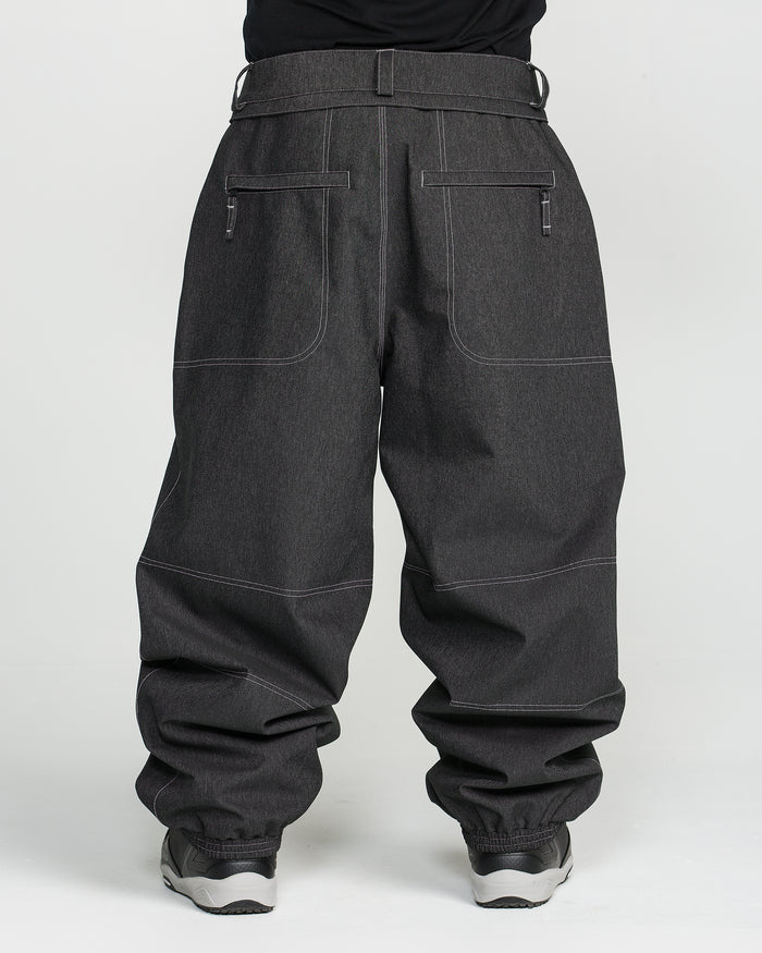 A person is wearing Volcom Men’s Lapper Pants in dark gray, featuring visible white stitching, large front pockets, and zippers, paired with gray sneakers and standing against a plain white background.