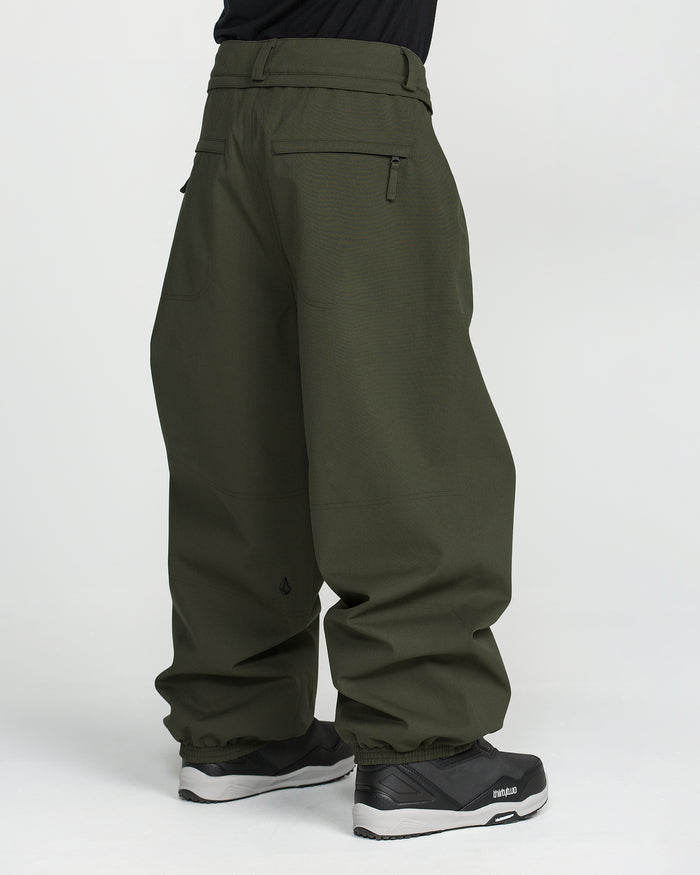 A person is shown from the side against a plain white background, wearing Volcom Men’s Lapper Pants in a loose-fitting, olive-green style with zippered back pockets and black sneakers. These baggy ski pants combine style and functionality for winter adventures.