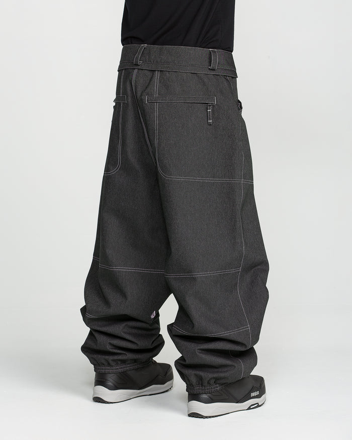 A person wearing Volcom Men’s Lapper Pants in dark gray, featuring white stitching, large pockets, and a loose, relaxed fit reminiscent of classic baggy snow pants, paired with black shoes and standing against a plain white background.