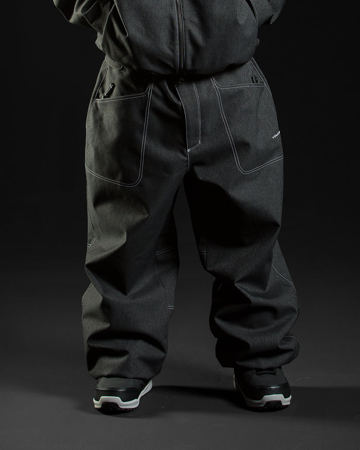 A person wearing Volcom Men’s Lapper Pants—loose-fitting, dark denim with white stitching and a PFAS-free DWR finish—pairs them with black sneakers while standing against a dark background. With hands in pockets and the upper body partially visible, the look channels the vibe of classic baggy snowboard pants.